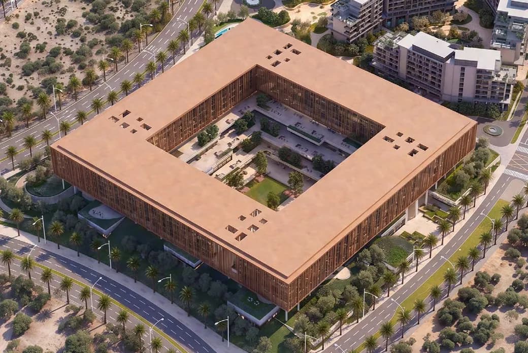Aerial view of a large square building with an open central courtyard, brown facade, surrounded by palm-lined streets and modern apartments.
