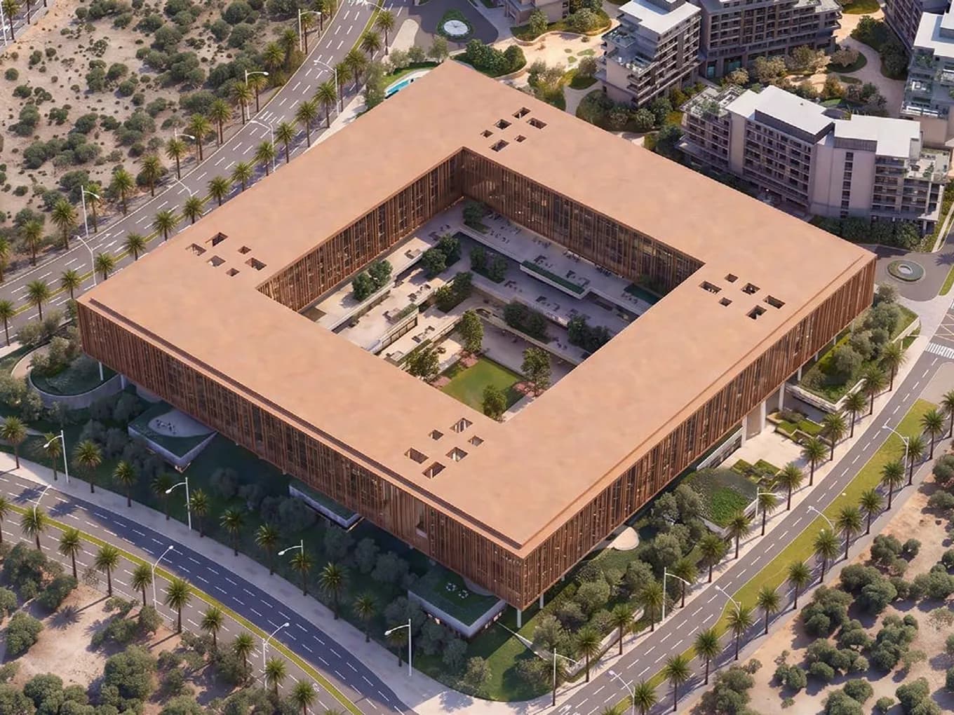 Aerial view of a large square building with an open central courtyard, brown facade, surrounded by palm-lined streets and modern apartments.