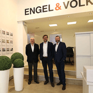 Drie mannen in pak staan in een Engel & Völkers makelaarskantoor. Vastgoedadvertenties hangen aan de muur.