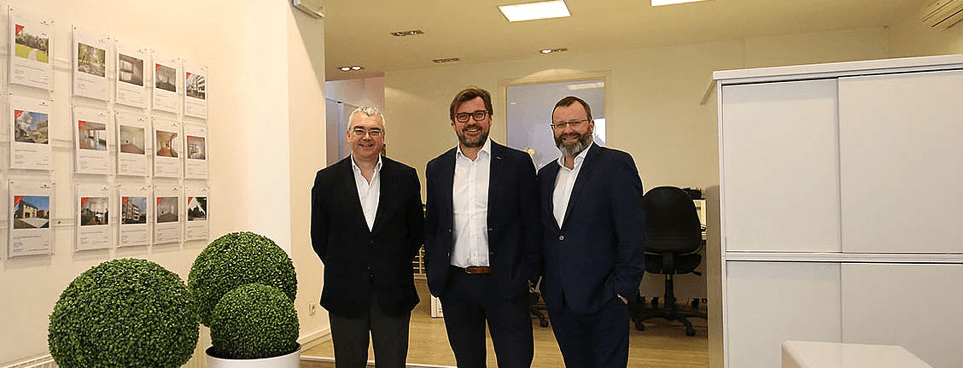 Drie mannen in pak staan in een Engel & Völkers makelaarskantoor. Vastgoedadvertenties hangen aan de muur.
