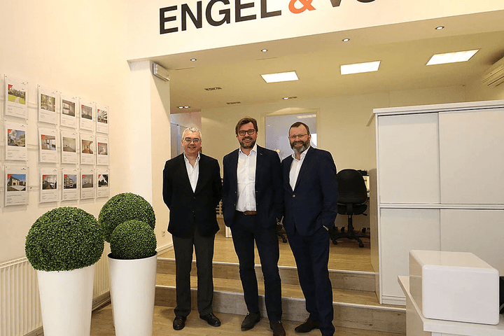 Trois hommes en costume se tiennent dans une agence immobilière Engel & Völkers. Des annonces immobilières sont accrochées au mur.