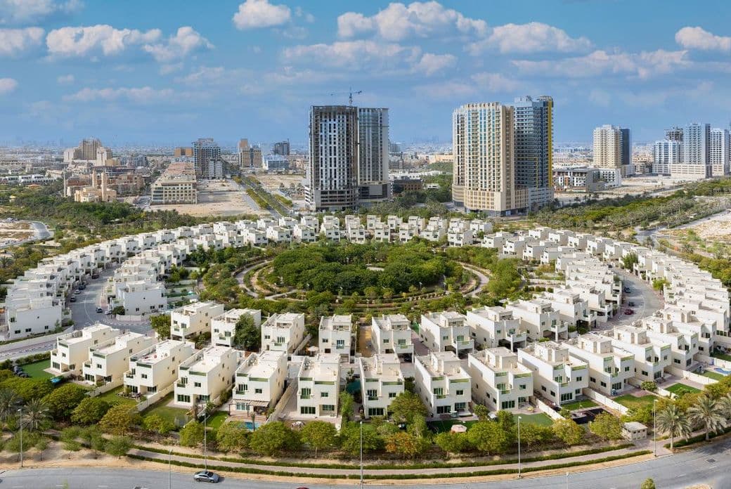 Circular residential complex with white houses and greenery, surrounded by roads, set against a backdrop of tall city buildings under a cloudy sky.