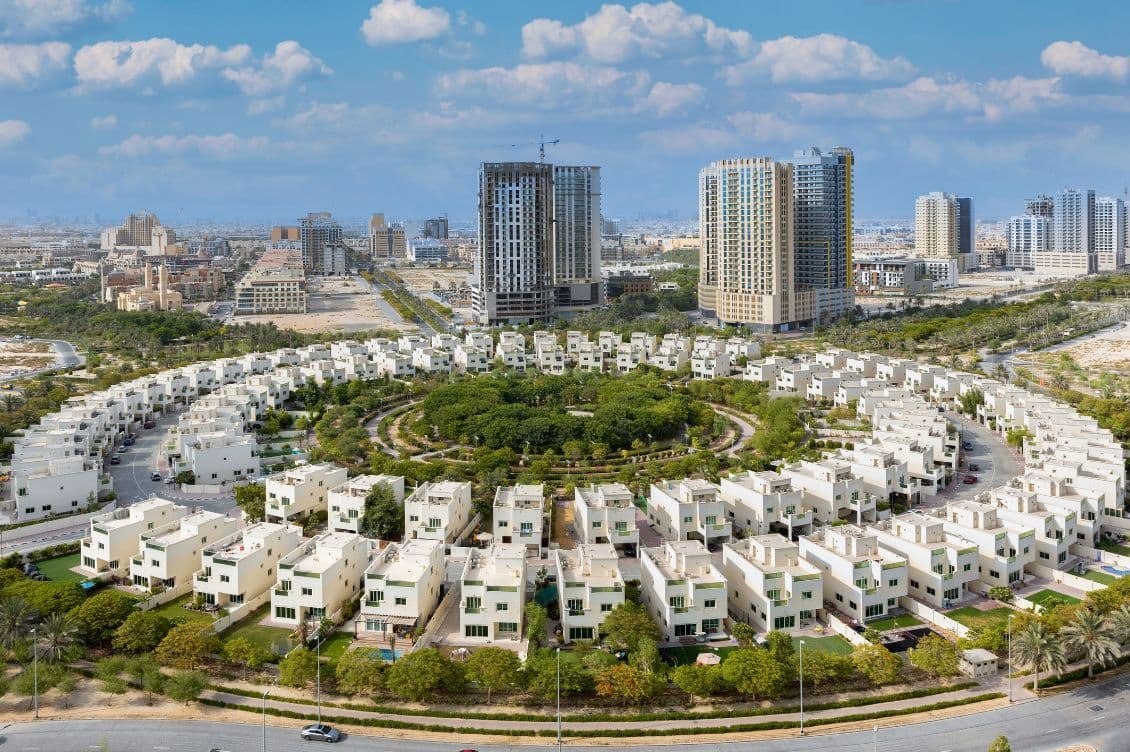 Aerial view of a circular residential complex with white houses, surrounded by greenery, and skyscrapers in the background under a partly cloudy sky.