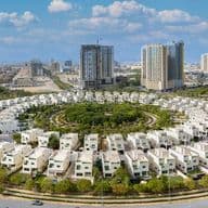 Aerial view of a circular neighborhood with white houses, green trees, and tall buildings in the background under a blue sky.