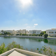 Waterfront view of luxury villas under a bright blue sky. Palm trees and green foliage frame the scene.