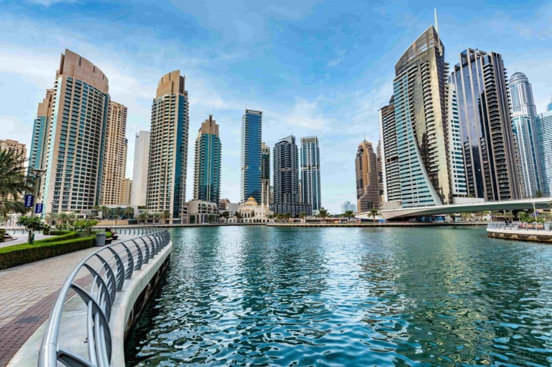 Skyline of modern skyscrapers along a waterfront promenade, with a clear blue sky and calm water reflecting the buildings.