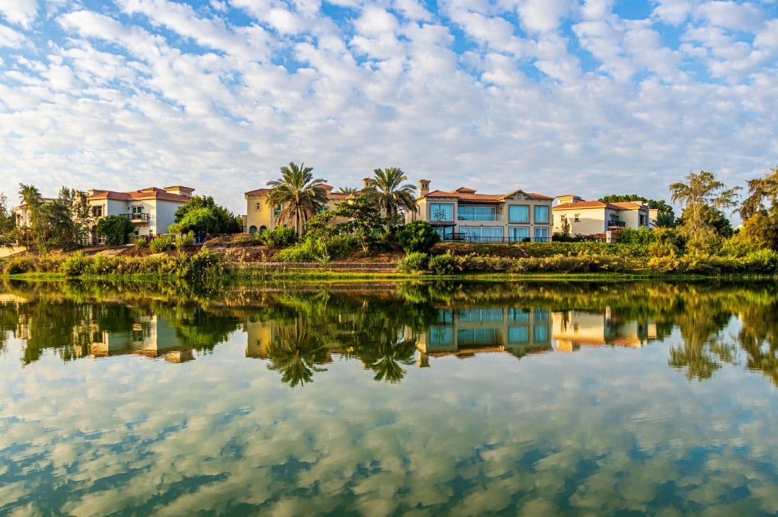 Luxurious houses with red-tiled roofs and palm trees reflected in a calm lake under a sky filled with scattered clouds.