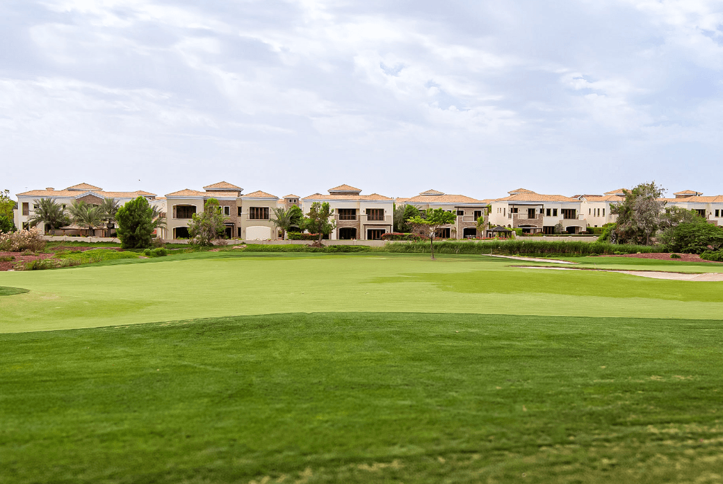 Eine Reihe beiger Villen mit orangen Dächern blickt unter einem bewölkten Himmel auf einen grünen Golfplatz.