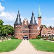 Blick auf das Holstentor, ein historisches Backsteintor mit zwei Türmen, umgeben von grünen Rasenflächen und Bäumen unter blauem Himmel in Lübeck, Deutschland.