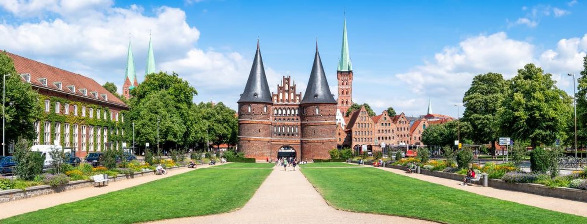 Wide view of the Holstentor, a historic brick gate with twin towers, surrounded by green lawns and trees under a blue sky in Lübeck, Germany.