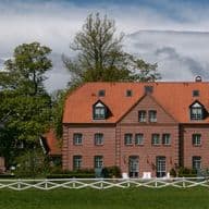 Ein großes Backsteinhaus mit rotem Dach, umgeben von üppigen grünen Bäumen und einem weißen Lattenzaun unter einem teilweise bewölkten Himmel.