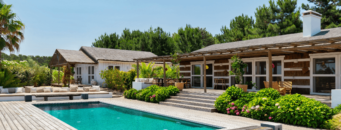 Uma villa luxuosa com um deck de madeira, piscina, área de estar ao ar livre e vegetação exuberante sob um céu azul claro.