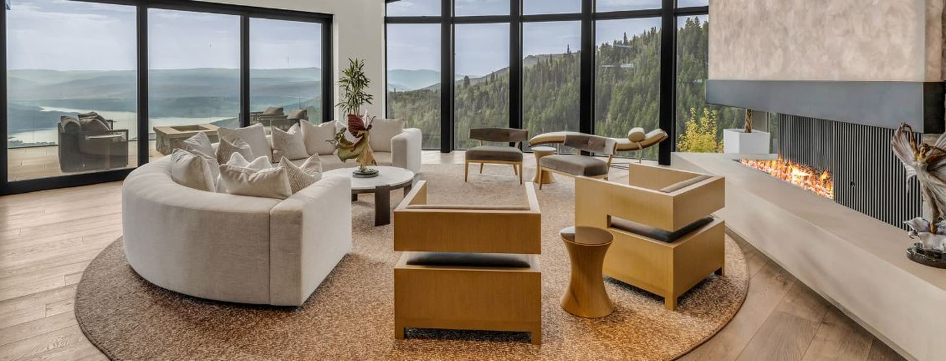 Luxurious living room with a curved sofa, modern chairs, fireplace, large windows overlooking mountains, and a round rug on wooden flooring.