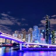 Illuminated cityscape at night featuring a modern bridge and tall skyscrapers reflecting on the water under a partly cloudy sky.
