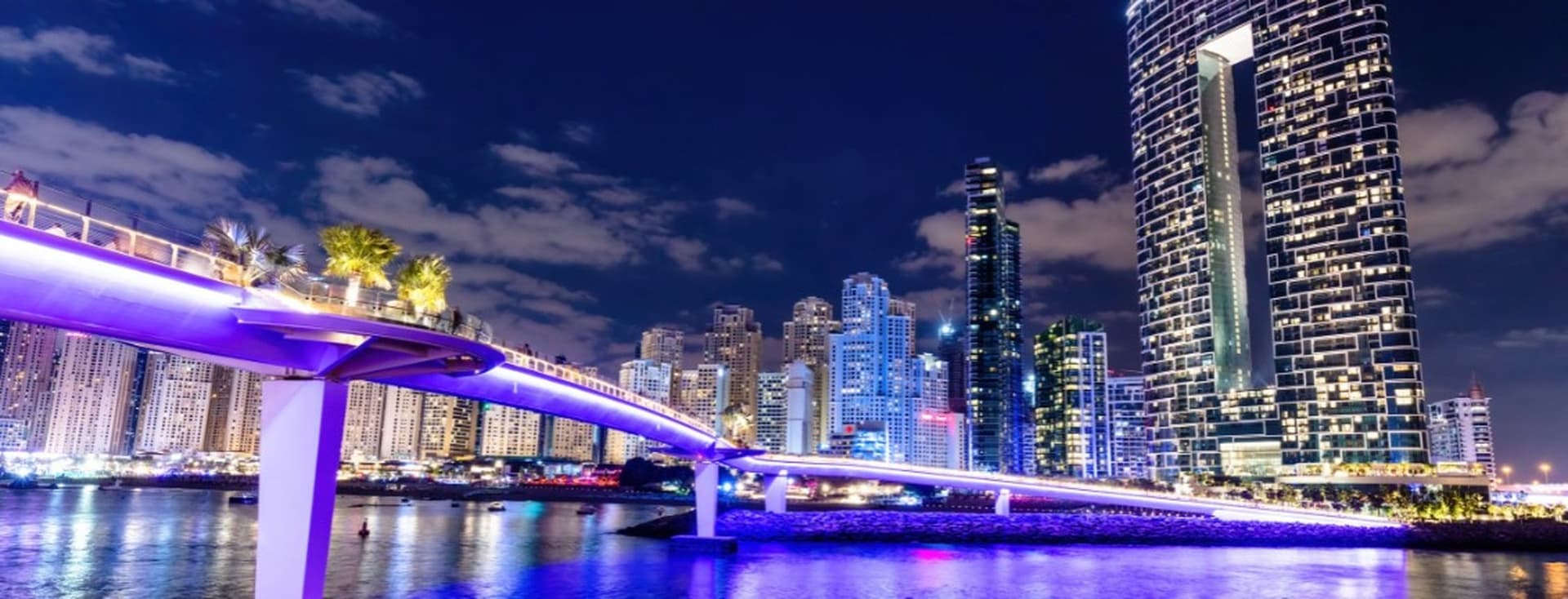 Illuminated cityscape at night featuring a modern bridge and tall skyscrapers reflecting on the water under a partly cloudy sky.