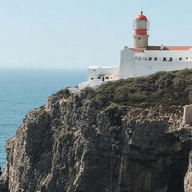 A white lighthouse with a red top sits atop a rocky cliff overlooking the ocean under a clear blue sky.