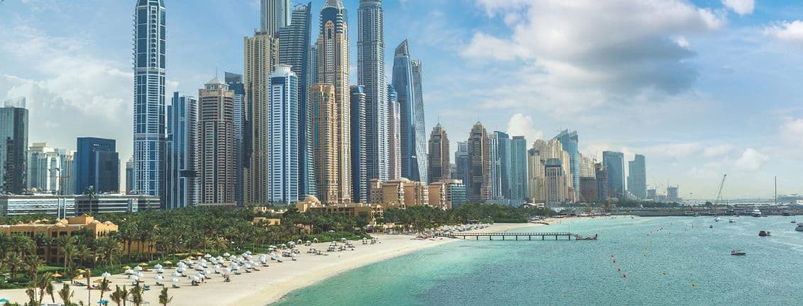 Skyline of tall, modern skyscrapers by a sandy beach and turquoise sea under a partly cloudy sky, with palm trees and beach umbrellas in the foreground.