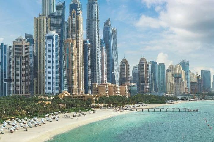Skyline of tall, modern skyscrapers by a sandy beach and turquoise sea under a partly cloudy sky, with palm trees and beach umbrellas in the foreground.