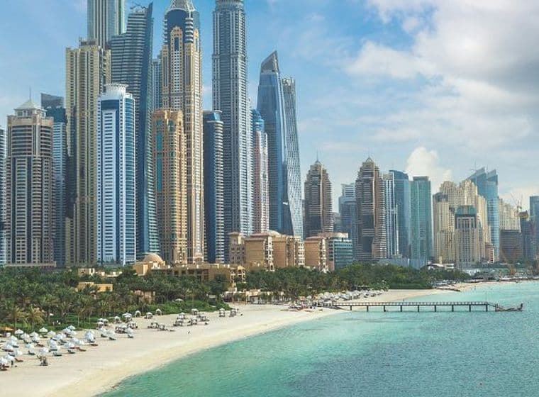 Skyline of tall, modern skyscrapers by a sandy beach and turquoise sea under a partly cloudy sky, with palm trees and beach umbrellas in the foreground.