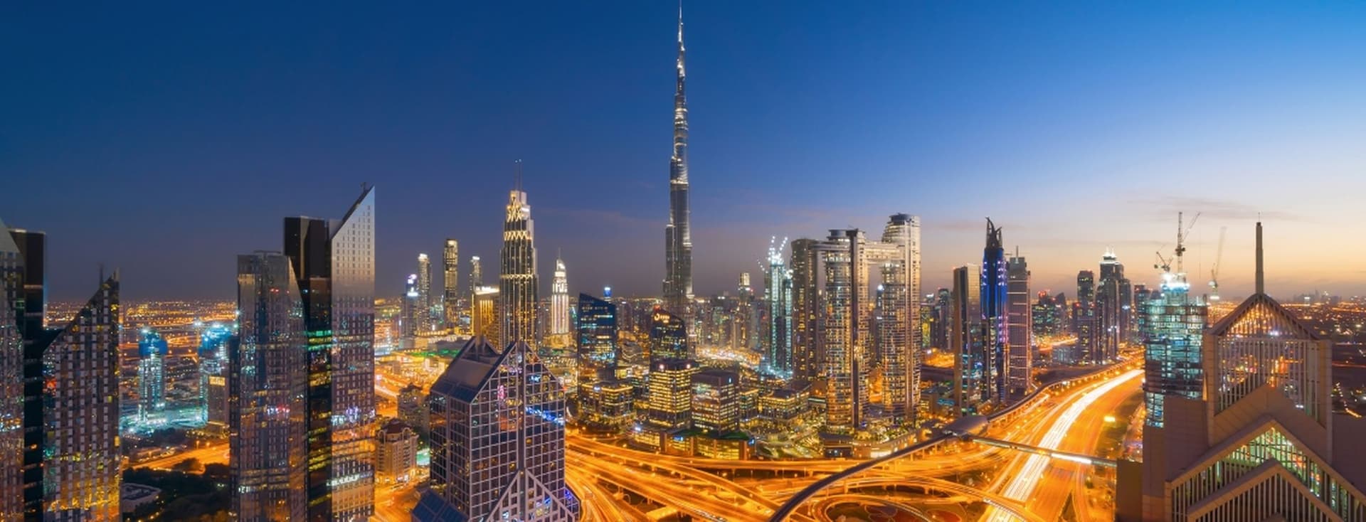 Aerial view of Downtown Dubai at dusk, featuring illuminated skyscrapers and winding highways under a deep blue sky.
