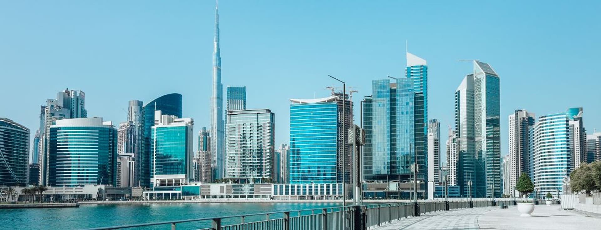 Dubai Skyline mit modernen blauen Glasgebäuden und dem Burj Khalifa unter einem klaren blauen Himmel, gesehen von einer Uferpromenade.