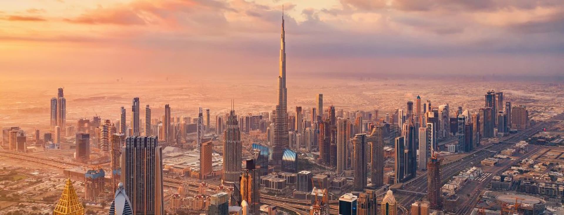 Aerial view of Downtown Dubai at sunset, featuring Burj Khalifa surrounded by numerous modern buildings under a partly cloudy sky.