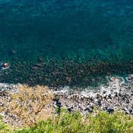 Primo piano di una costa rocciosa siciliana. Pietre laviche di diverse dimensioni si alternano lungo la linea di costa, bagnate dalle onde del mar Mediterraneo. L'acqua, di un blu profondo che sfuma in tonalità di verde acqua, lambisce i massi. Vegetazione mediterranea con cespugli verdi occupa la parte superiore dell'immagine, creando un contrasto con la durezza delle rocce sottostanti. La scena cattura l'essenza selvaggia e suggestiva del paesaggio costiero siciliano.