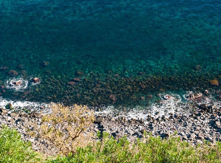 Primo piano di una costa rocciosa siciliana. Pietre laviche di diverse dimensioni si alternano lungo la linea di costa, bagnate dalle onde del mar Mediterraneo. L'acqua, di un blu profondo che sfuma in tonalità di verde acqua, lambisce i massi. Vegetazione mediterranea con cespugli verdi occupa la parte superiore dell'immagine, creando un contrasto con la durezza delle rocce sottostanti. La scena cattura l'essenza selvaggia e suggestiva del paesaggio costiero siciliano.