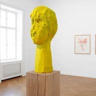 Sala espositiva luminosa con scultura in legno giallo su piedistallo in parquet e tre disegni su pareti bianche