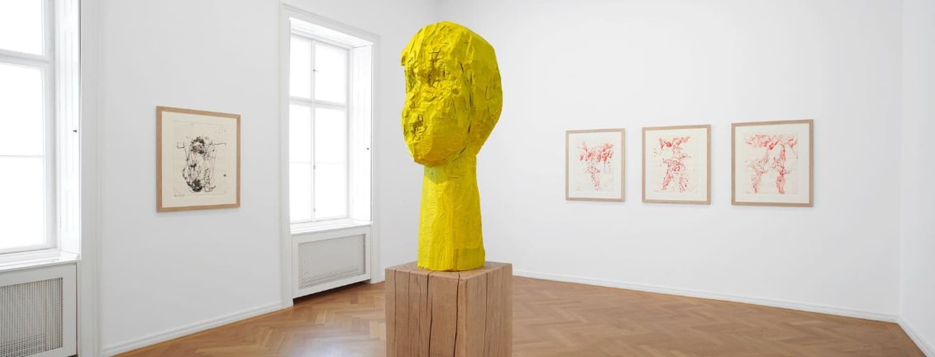 Heller Galerieraum mit gelber Holzskulptur auf Sockel Parkett und drei Zeichnungen an weißen Wänden