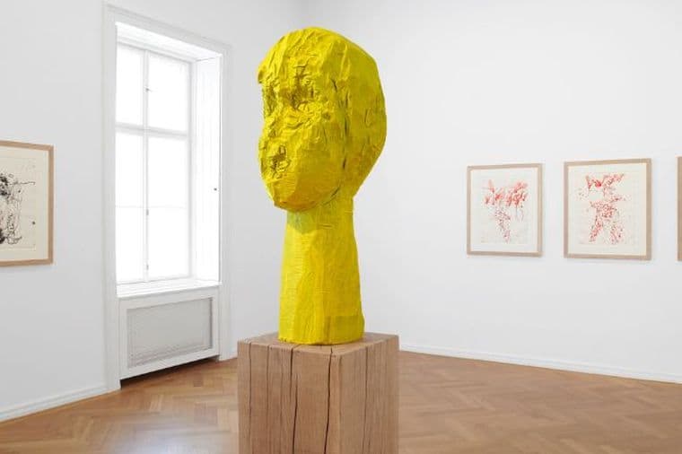 Bright gallery space with yellow wooden sculpture on a pedestal, parquet flooring and three drawings on white walls