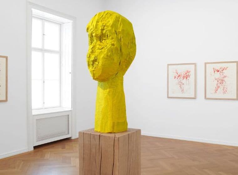 Bright gallery space with yellow wooden sculpture on a pedestal, parquet flooring and three drawings on white walls