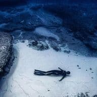 Ein Freitaucher im Neoprenanzug liegt auf dem sandigen Meeresgrund zwischen Felsformationen im tiefblauen Wasser.