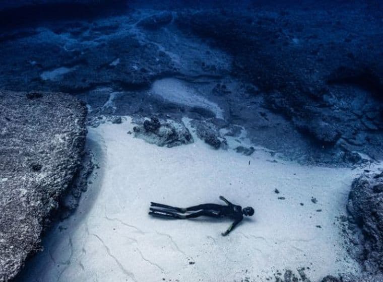 Underwater scene: a small dark figure lying on white sand between two rock formations.