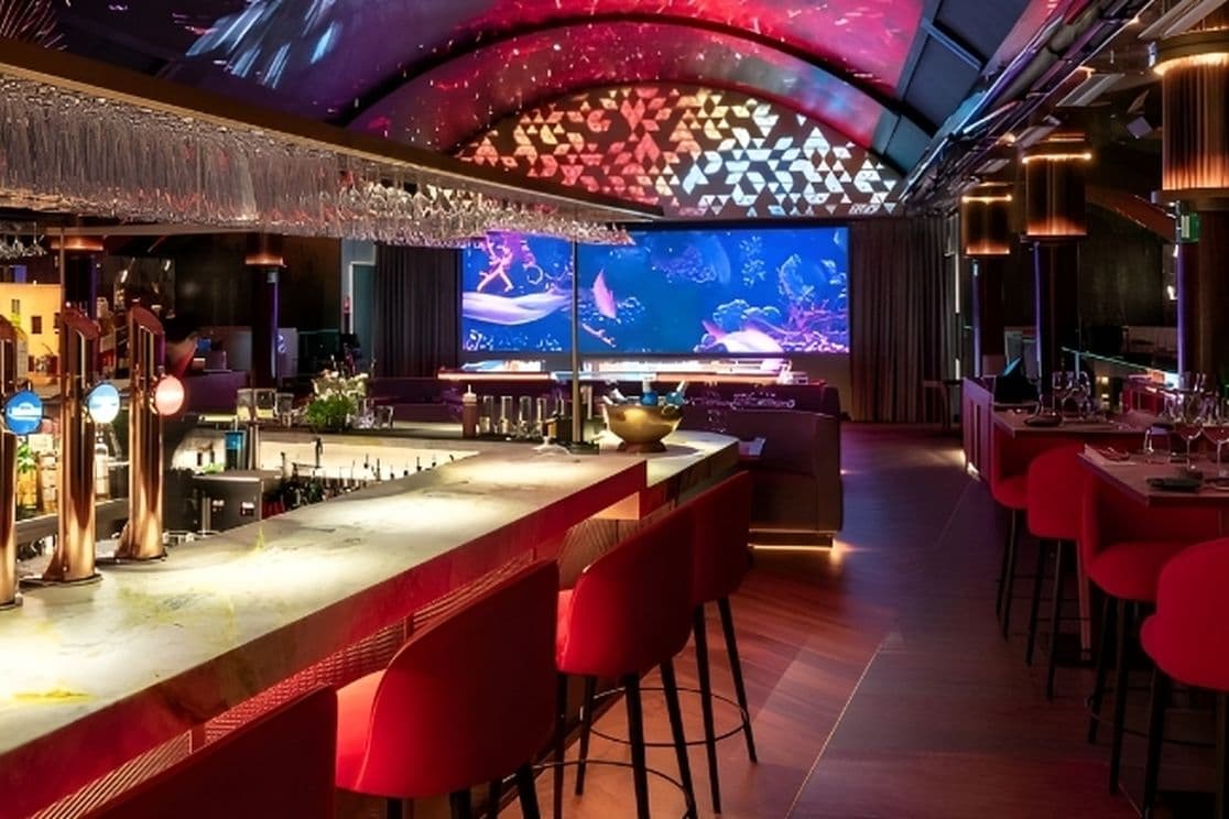 Modern bar and lounge with illuminated counter, red seats, and ambient lighting. Large screen in the background displays a colorful image.