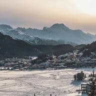Schneebedeckte Landschaft mit einer Stadt, die sich in ein Tal schmiegt, umgeben von Bergen unter der untergehenden Sonne. Auf dem zugefrorenen See sind Skispuren zu sehen.