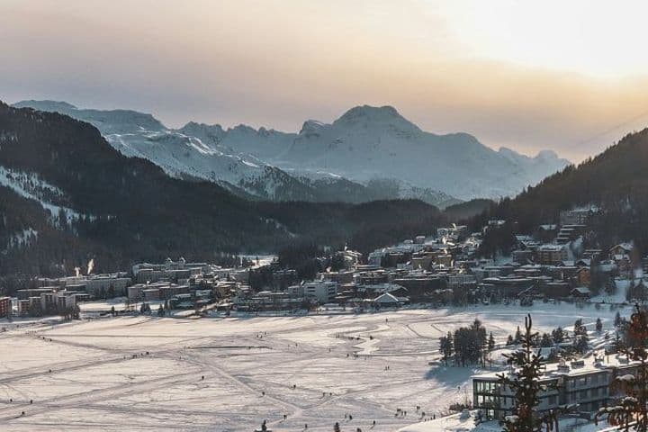 Schneebedeckte Landschaft mit einer Stadt, die sich in ein Tal schmiegt, umgeben von Bergen unter der untergehenden Sonne. Auf dem zugefrorenen See sind Skispuren zu sehen.