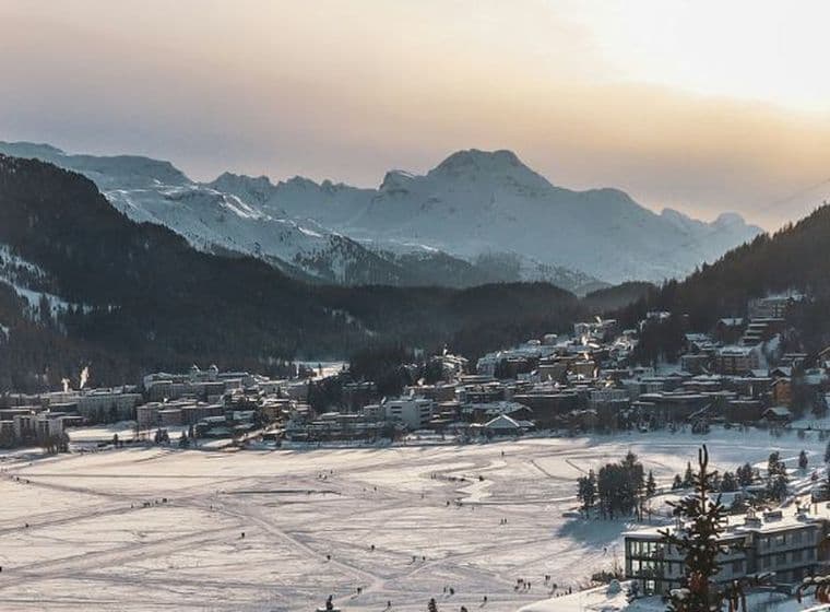 Paesaggio innevato con una città adagiata in una valle, circondata da montagne sotto il sole al tramonto. Sul lago ghiacciato si vedono tracce di sci.