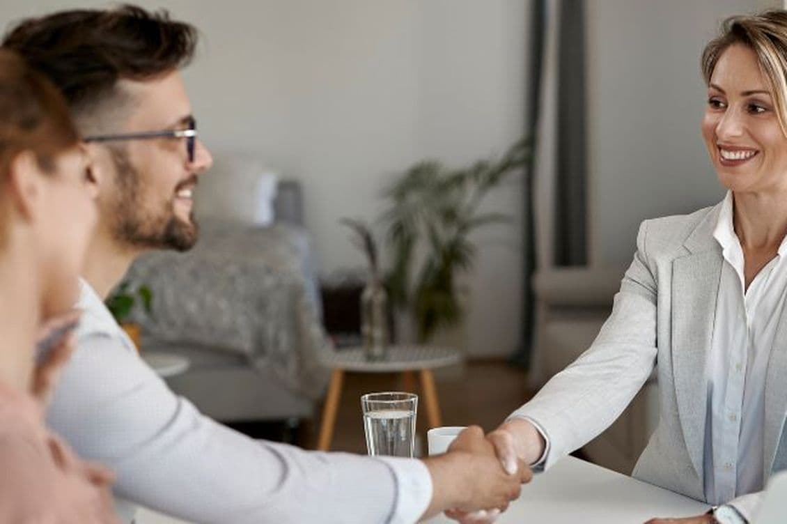 A smiling professional mortgage broker in a suit shakes hands with a seated man and woman in an office setting.