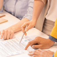 People collaborating at a table, pointing at documents and discussing ideas, with a coffee cup and plant nearby.