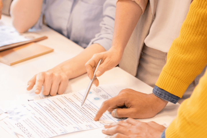 Mehrere Personen arbeiten gemeinsam an einem Schreibtisch, zeigen auf Dokumente mit Diagrammen und Grafiken; eine Kaffeetasse und ein Klemmbrett liegen daneben.