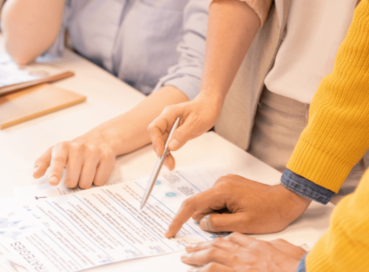 Mehrere Personen arbeiten gemeinsam an einem Schreibtisch, zeigen auf Dokumente mit Diagrammen und Grafiken; eine Kaffeetasse und ein Klemmbrett liegen daneben.