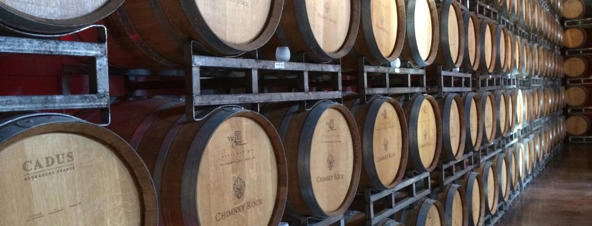 Rows of stacked wooden wine barrels in a dimly lit cellar, each marked with different winery labels.