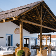 A modern patio with a thatched roof, wicker furniture, and a pool, adjacent to a white house under a clear blue sky.