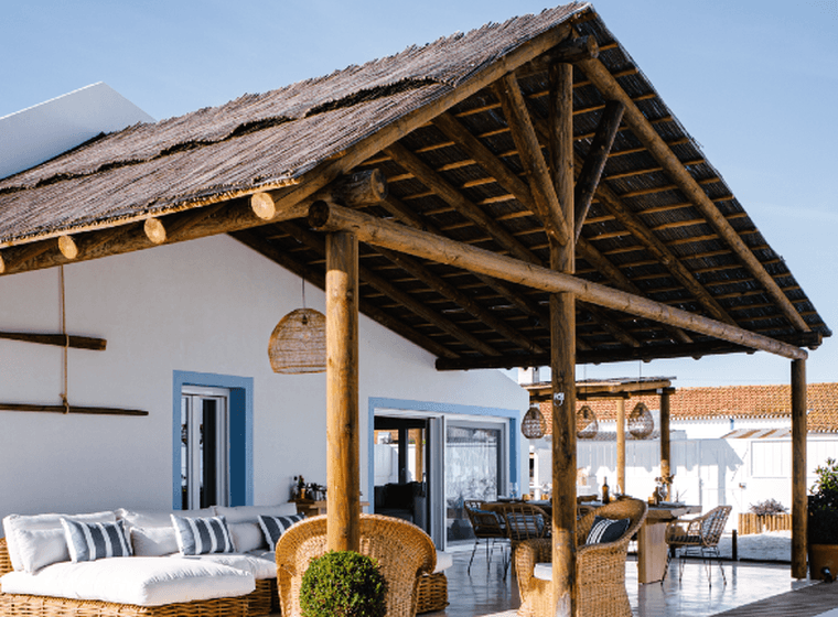 Eine moderne Terrasse mit Reetdach, Korbmöbeln und einem Pool, angrenzend an ein weißes Haus unter einem klaren blauen Himmel.