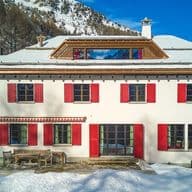 Large white Alpine house with red shutters and a terrace, surrounded by snow-covered fir trees and mountains under a bright blue sky.