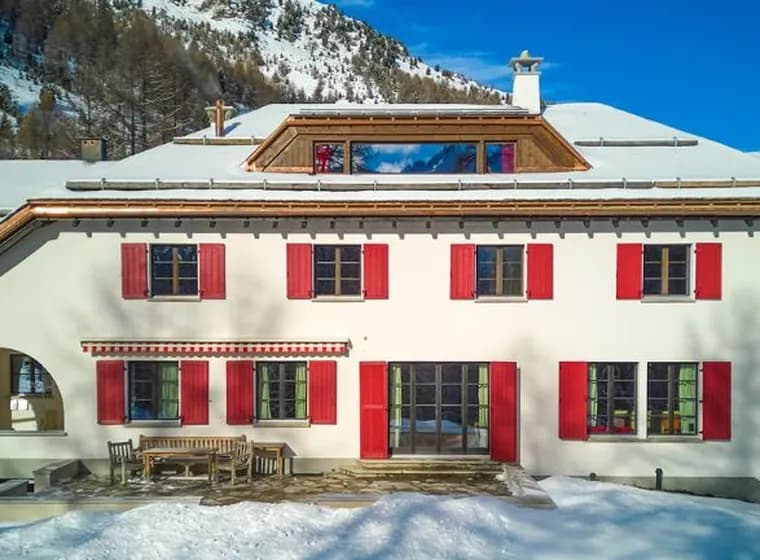 Large white Alpine house with red shutters and a terrace, surrounded by snow-covered fir trees and mountains under a bright blue sky.