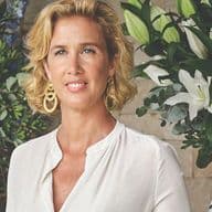 Κομψή γυναίκα με ανοιχτόχρωμο φόρεμα δίπλα σε τραπέζι με μεγάλες ανθοσυνθέσεις σε διακοσμητικά βάζα