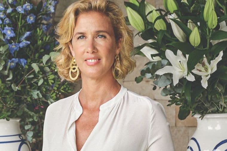 Κομψή γυναίκα με ανοιχτόχρωμο φόρεμα δίπλα σε τραπέζι με μεγάλες ανθοσυνθέσεις σε διακοσμητικά βάζα