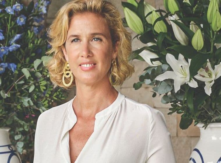 Κομψή γυναίκα με ανοιχτόχρωμο φόρεμα δίπλα σε τραπέζι με μεγάλες ανθοσυνθέσεις σε διακοσμητικά βάζα